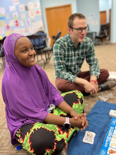 A smiling girl wearing a hijab is in the foreground and a smiling teacher wearing glasses is in the background.